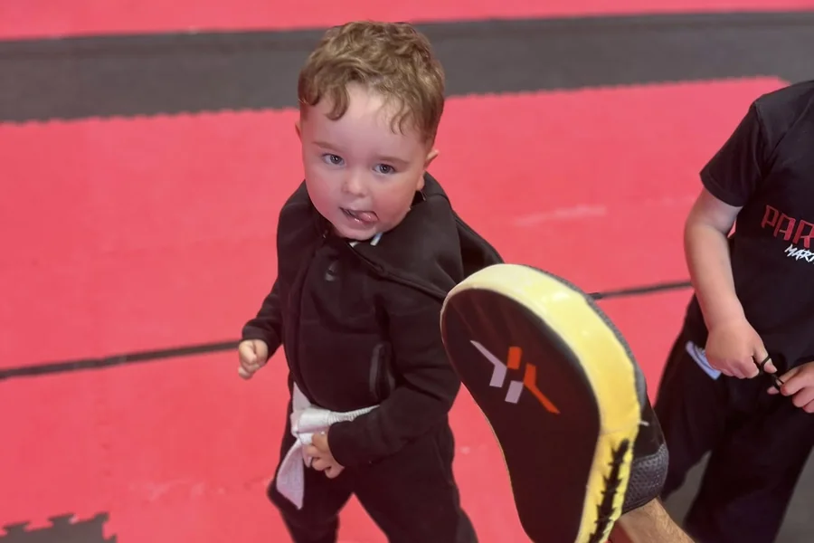 A Little Ninja in a white belt putting a punch into a coach's focus pad
